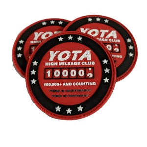 May include: Three red and black round patches with white text and stars. The center patch reads "YOTA HIGH MILEAGE CLUB 100002 100,000+ AND COUNTING PRIDE IN MAINTENANCE PRIDE IN OWNERSHIP".
