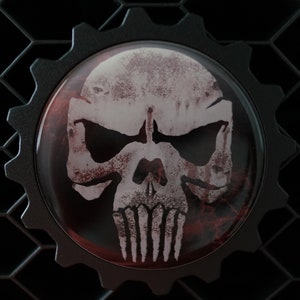 May include: A black gear-shaped emblem with a white skull and crossbones design. The skull has a distressed, weathered look and is set against a red background.