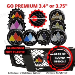 May include: A set of 3.4 inch or 3.75 inch aluminum car emblems with a mountain range design. The emblems are available in a variety of colors, including red, orange, yellow, blue, white, gray, and black. The emblems are also available in a gear or round shape. The text "GO PREMIUM" is displayed at the top of the image. The text "ALUMINUM NOT PLASTIC" is displayed at the bottom of the image. The text "Grille Mount or Flat Mount Options!" is displayed at the bottom left of the image. The text "Have Fun, Be Different!" is displayed at the bottom right of the image.