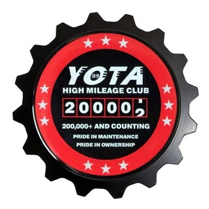 May include: A black, gear-shaped emblem with a red and black circular center. The center features the text "YOTA HIGH MILEAGE CLUB" and "20000,". Below, it reads "200,000+ AND COUNTING, PRIDE IN MAINTENANCE, PRIDE IN OWNERSHIP". White stars encircle the center.