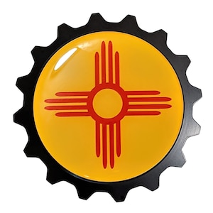May include: A round, black-edged emblem featuring the New Mexico state flag. The flag design includes a yellow background with a red Zia sun symbol at the center. The emblem has a glossy finish.