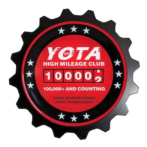 May include: A black and red metal gear-shaped sign with the text "YOTA HIGH MILEAGE CLUB" and a digital display reading "10000". The sign also includes the text "100,000+ AND COUNTING", "PRIDE IN MAINTENANCE", and "PRIDE IN OWNERSHIP".