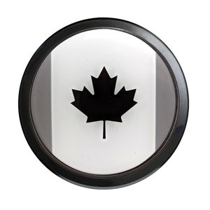 May include: A black and white Canadian flag with a red maple leaf in the center, displayed in a black circular frame.