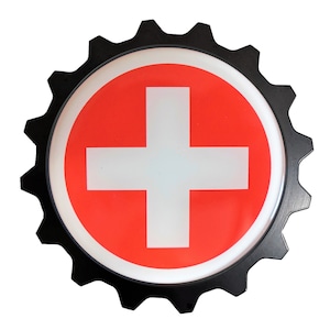 May include: A round, black-edged sign featuring the Swiss flag. The sign has a red background with a white cross in the center. The outer edge is shaped like a gear.