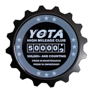 May include: A black and silver gear-shaped emblem with the text "YOTA HIGH MILEAGE CLUB" and a digital odometer reading "500002". The text "500,000+ AND COUNTING" is below the odometer. The text "PRIDE IN MAINTENANCE" and "PRIDE IN OWNERSHIP" are below the counting text.