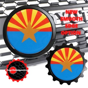 May include: Two black and gold Arizona state flag car emblems. The emblem on the left has a smooth edge. The emblem on the right has a gear-shaped edge. Both emblems feature the Arizona state flag with a red sun, yellow rays, and a gold star on a blue background. The text "NEW SMOOTH EDGE OPTION" is visible in the background.
