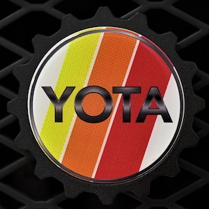 May include: A black and white emblem with a gear-shaped border. The center of the emblem features a red, orange, and yellow striped design with the word "YOTA" in black letters.