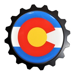 May include: A black gear-shaped emblem with a blue, white, red, and yellow Colorado state flag design.
