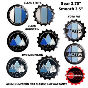 May include: A set of six different car emblems with various designs. The emblems are made of aluminum and resin and feature a 4WD Off-Road Explorer design, a mountain range design, and a YOTA design. The emblems are available in two sizes: 3.75 inches and 3.5 inches. The text "Gear 3.75" and "Smooth 3.5" is displayed above the emblems. The text "YOTA FAT" is displayed above the YOTA emblem. The text "CLEAN STRIPE" is displayed above the 4WD Off-Road Explorer emblem. The text "CLEAN MOUNTAIN" is displayed above the mountain range emblem. The text "4WD MOUNTAIN" is displayed below the mountain range emblem. The text "ALUMINUM/RESIN NOT PLASTIC 1-YR WARRANTY" is displayed below the emblems. The text "thegrillebadgestore.com" is displayed below the emblems.