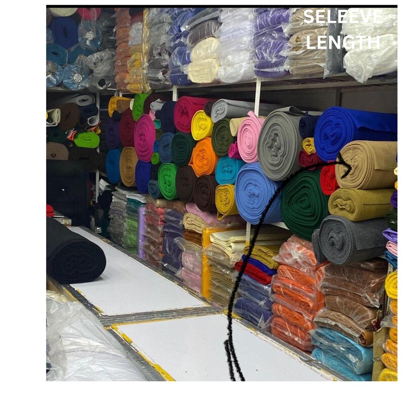 Op de afbeelding: Schappen vol met rollen stof in verschillende kleuren, waaronder roze, blauw, groen en oranje. De stoffen rollen zijn netjes opgestapeld, sommige verpakt in plastic. De tekst "SLEEVE LENGTH" is zichtbaar bovenaan.