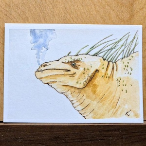 May include: Watercolor painting of a yellow and brown dragon with green spikes and a wisp of blue smoke. The dragon is facing left, with a detailed textured skin and a thoughtful expression. The artwork is on a white rectangular card.