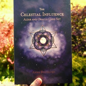 May include: A rectangular box for The Celestial Influence Altar and Oracle Card Set by Anna Bousquet. The cover features a dark purple cosmic background with a central geometric star design containing a moon illustration and white star accents.