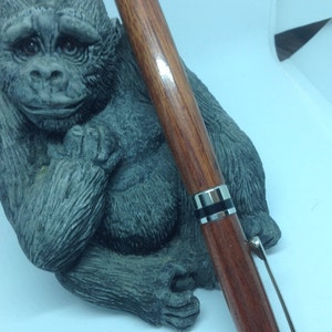 Könnte beinhalten: Ein Holzstift mit einem silbernen Band und schwarzen Akzenten. Der Stift liegt auf einer grauen, geformten Gorilla-Figur.
