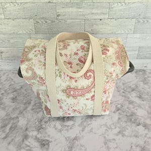 May include: A fabric tote bag featuring a cream-colored base with a repeating floral and paisley pattern in shades of pink, red, and muted green. The bag includes two thick, beige woven cotton handles attached to the front and back panels.