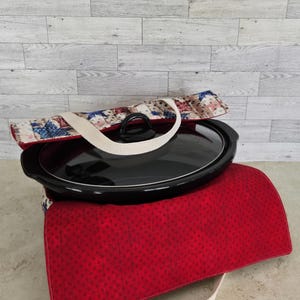 May include: A black oval slow cooker with a glass lid sits inside a quilted fabric carrier. The carrier features a red fabric with small dark dots on the bottom and a top flap patterned with blue and red stars on a light background.