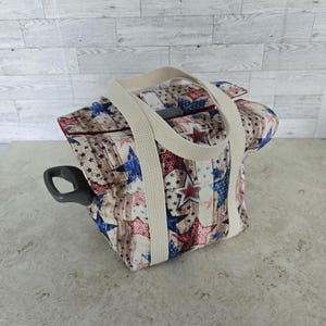 May include: A patriotic-themed tote bag featuring a pattern of red, white, and blue stars on a wood-grain background. The bag has a cream-colored handle and a dark gray handle on the side.
