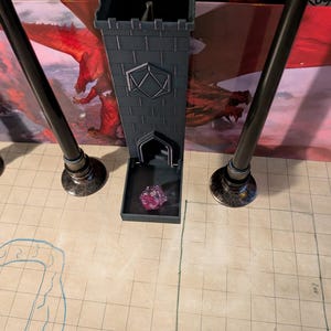 May include: A black plastic tower with a square base and a small opening at the top. The tower has a decorative design on the front and a small compartment at the bottom. A pink and purple die is inside the compartment.