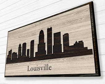May include: Wooden wall art featuring the Louisville skyline in a dark brown silhouette against a light wood grain background. The word "Louisville" is printed in a simple font at the bottom of the artwork. The piece is framed in a dark brown border.