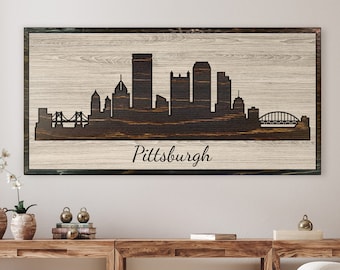 May include: A wood sign with a black silhouette of the Pittsburgh skyline and the word "Pittsburgh" written below. The sign has a light wood background with dark wood grain.