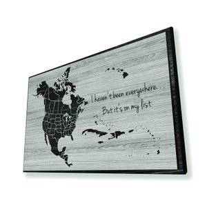 May include: A wood-grain textured wall art piece with a black frame. The art features a map of North America with the text "I haven't been everywhere... But it's on my list."