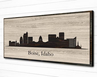 May include: Wooden wall art featuring the Boise, Idaho skyline silhouette. The artwork is a dark brown against a light wood grain background, framed in a darker wood. The city name is printed in black.