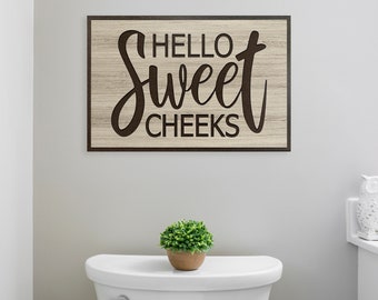 May include: A wooden sign with a brown background and black text that reads "Hello Sweet Cheeks". The sign is hanging on a white wall above a white toilet.