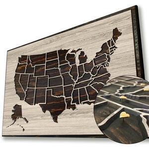 May include: A wooden map of the United States with a dark brown wood grain finish. The map is mounted on a light wood frame. The map is designed to be used as a push pin map, with small gold heart-shaped push pins placed in the map.