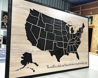 May include: Wooden wall art featuring a map of the United States with individual states outlined in black against a light wood grain background. The artwork is framed in black and includes the text: "The world is a book, and those who do not travel read only a page."