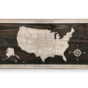 May include: A wooden map of the United States with a white outline of the states and a compass in the lower right corner. The text "Johnson Family Travels" is at the top of the map and "Been There, Pinned That." is at the bottom.