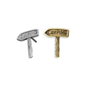 May include: Two metal camping sign pins, one silver and one gold, with the word "CAMPING" on the sign.