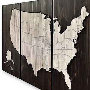 May include: A three-panel wooden wall art piece featuring a raised, white outline map of the United States. The map is divided into states and includes Alaska and Hawaii. The wood is a dark brown color.