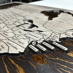 May include: A wooden map of the United States with the states carved out and labeled. The map is stained a dark brown color and has a light gray background. The states of New York, Pennsylvania, Virginia, North Carolina, Maryland, Delaware, New Jersey, Rhode Island, Maine, Vermont, New Hampshire, Massachusetts, and Connecticut are visible.