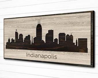 May include: A wood sign with a black silhouette of the Indianapolis skyline. The sign has a light brown wood grain and the text "Indianapolis" is printed in black below the skyline.