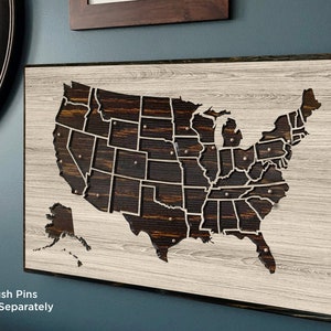 May include: A wooden map of the United States with each state outlined. The map is on a whitewashed wood background with a dark wood frame. The text "Push Pins Sold Separately" is at the bottom of the image.