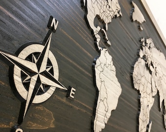 May include: A wooden world map with a compass rose in the lower left corner. The map is painted in shades of brown and white, with the continents outlined in white. The compass rose is also painted in white, with the letters N, S, E, and W.
