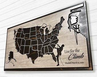 May include: Wooden wall art featuring a map of the United States with state outlines. The artwork includes silhouettes of rock climbers and the text "Live for the Climb" with a date. The piece has a dark brown border and a light wood grain background.