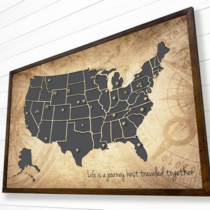 May include: A framed map of the United States with a dark gray outline of the states and gold push pins. The map has an aged, vintage look with a compass rose and the text "Life is a journey best traveled together".