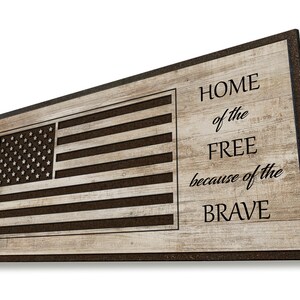 May include: A wooden doormat with a brown woodgrain pattern and a black border. The doormat features a cutout of the American flag and the text "HOME of the FREE because of the BRAVE".