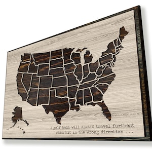 May include: A wooden wall art piece featuring a laser-cut map of the United States with each state outlined. The map is on a light wood background with a dark wood frame. The text "A golf ball will ALWAYS travel furthest when hit in the wrong direction..." is printed below the map.