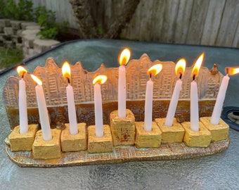 May include: A gold menorah with nine white candles lit, each with a small flame. The menorah is decorated with a textured design and is sitting on a table.
