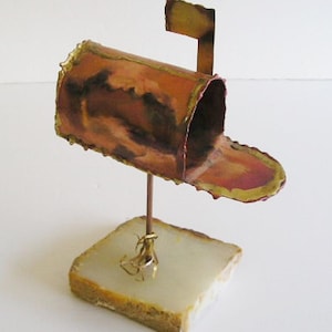 May include: A small, copper-colored mailbox sculpture with a flag on top, mounted on a white marble base.