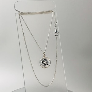 May include: A silver necklace with a round pendant featuring a black and white floral design. The necklace is displayed on a clear acrylic stand, showcasing the delicate chain and clasp.