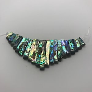 May include: A curved arrangement of abalone shell beads. The rectangular beads exhibit iridescent colours, including green, blue, and purple. They are strung on a clear string, ideal for crafting jewellery. Each bead is approximately 1 cm in length.