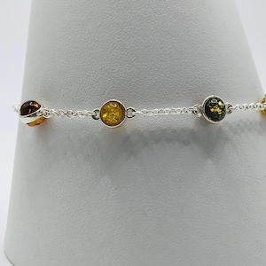 May include: A silver bracelet with five amber beads in shades of yellow, brown, and green. The round amber stones are set in silver bezels and linked by a delicate silver chain. The bracelet is displayed on a white surface.