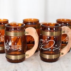 May include: Set of six amber-colored glass beer mugs with wooden handles and brass accents. Each mug features a unique white etched design, including a wagon wheel, campfire, and cowboy-themed illustrations. A vintage barware set.