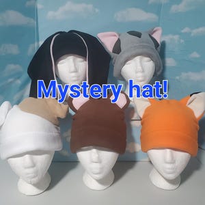 May include: A collection of fleece hats in various animal designs, including a black and white panda, a gray elephant, a white dog, a brown bear, and an orange fox. The text "Mystery hat!" is displayed in blue.