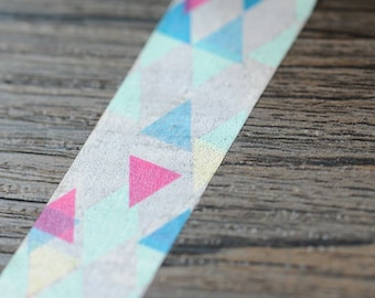 May include: Decorative washi tape with a geometric pattern of pink, blue, and light green triangles on a white background. The tape is partially rolled, revealing the pattern. The tape is on a wooden surface.