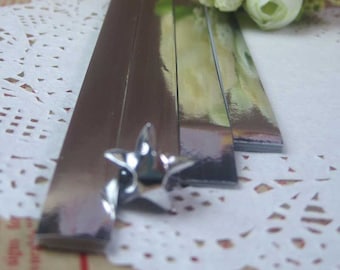 Puede incluir: Un conjunto de cinco tiras de papel metálico plateado, cada una con una superficie brillante y reflectante. Las tiras están dispuestas en una pila, con la tira superior ligeramente desplazada de las demás. Las tiras están sobre una puntilla blanca.