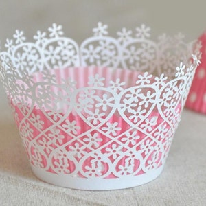 May include: White cupcake liners with a delicate lace design featuring a heart pattern. The liners are pink on the inside.
