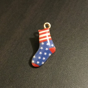 May include: A small, gold-coloured charm in the shape of a sock with an American flag design. The sock is red, white, and blue with stars and stripes.